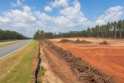 Multi-acre commercial site being cleared beside a four-lane road, machines working among mulch windrows with silt fence installed