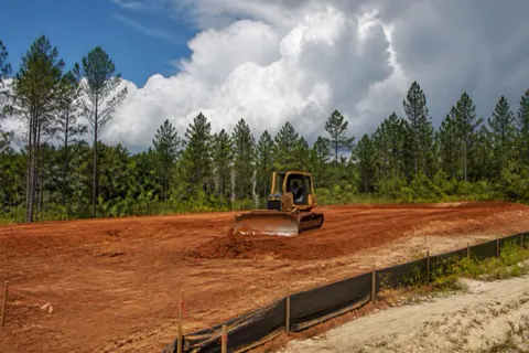 Compact dozer shaping rust-red Georgia clay into a homesite pad, string lines set and thunderheads building behind the pine line